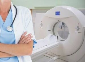 immagine in primo piano di metà busto di una dottoressa a braccia conserte con camice e stetofonendoscopio al collo non si vede però il volto sullo sfondo un macchinario per la risonanza magnetica